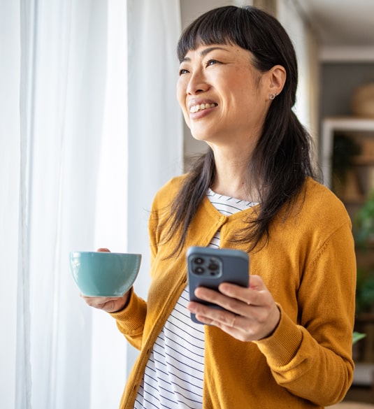 Smiling woman holding a coffee cup and smart phone, looking out a window on a sunny day. Smiling woman holding a coffee cup and smart phone, looking out a window on a sunny day.